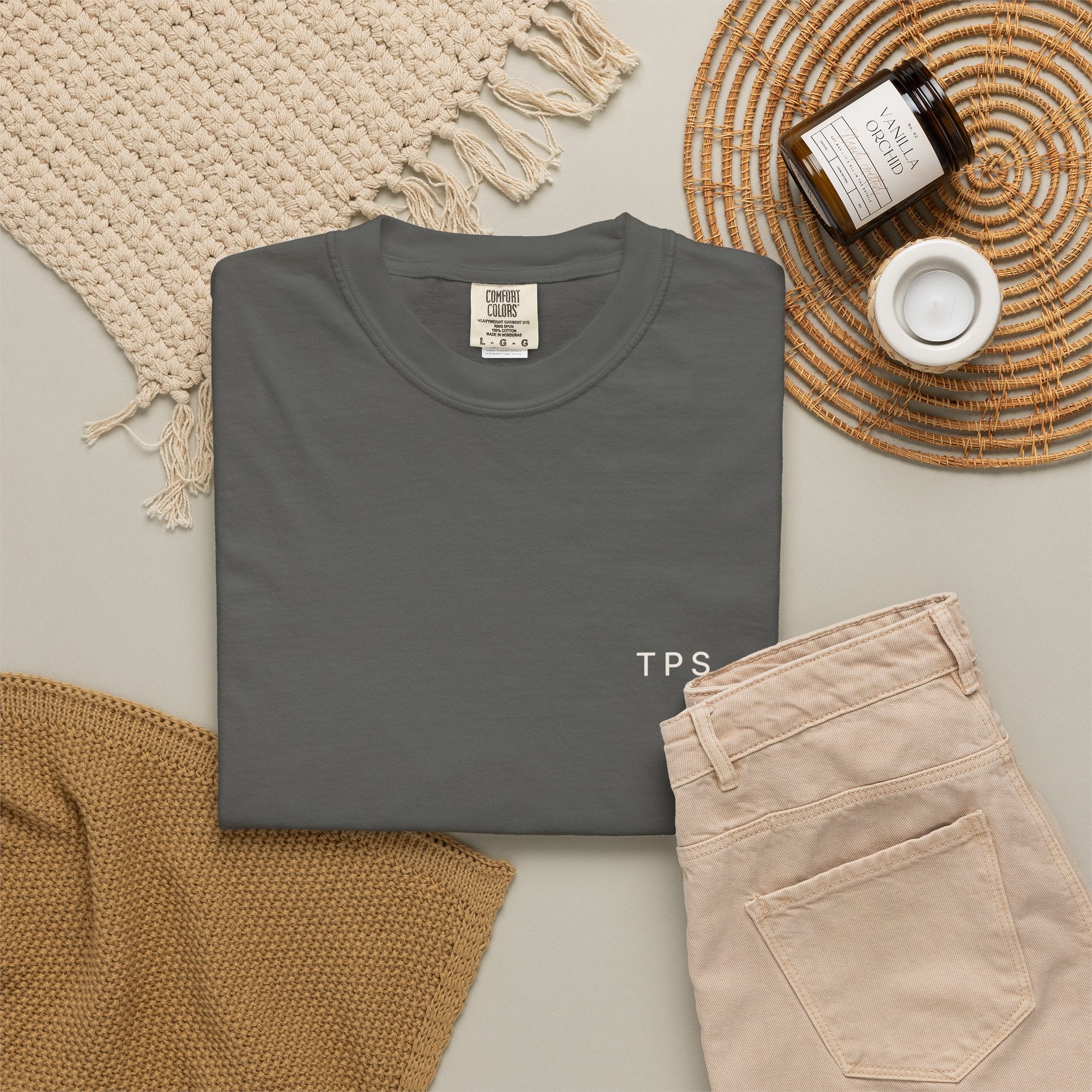 Folded gray t-shirt with 'TPS' text on a neutral surface with beige pants and decorative items.